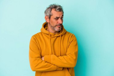 Orta yaşlı beyaz bir adam mavi arka planda izole edilmiş. Kameraya alaycı bir ifadeyle bakıyor..