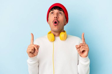 Genç melez adam mavi arka planda izole edilmiş müzik dinleyip ağzı açık bir şekilde üst tarafı işaret ediyor..