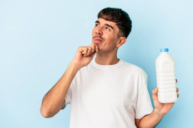 Mavi arka planda bir şişe süt tutan melez genç adam şüpheli ve şüpheci bir ifadeyle yana bakıyor..