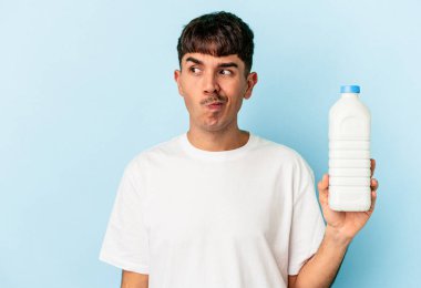 Mavi arka planda izole edilmiş bir şişe sütü tutan melez genç adam kafası karışmış ve kendinden emin değil..