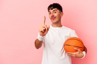 Genç melez adam pembe arka planda izole bir şekilde basketbol oynuyor. Parmağıyla bir numarayı gösteriyor..
