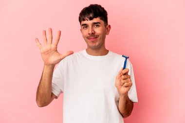 Genç melez adam pembe arka planda izole edilmiş jilet tutuyor. Gülümsüyor beş numarayı parmaklarıyla gösteriyor..