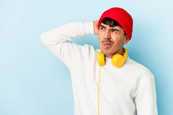 Young mixed race man listening to music isolated on blue background touching back of head, thinking and making a choice.