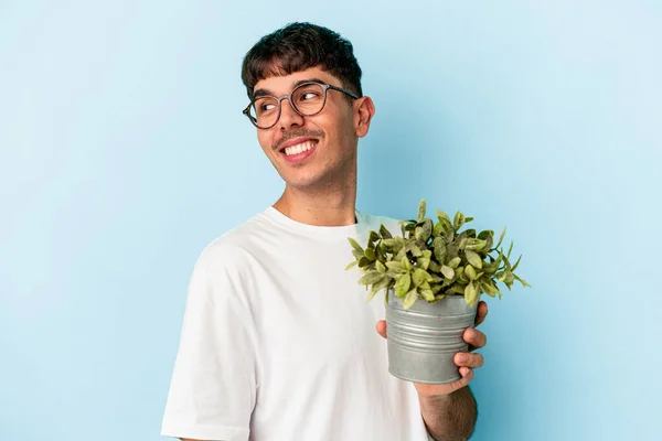 Młody Mieszane Wyścigu Człowiek Gospodarstwa Roślin Odizolowanych Niebieskim Tle Patrzy — Zdjęcie stockowe