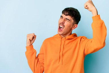 Genç melez adam mavi arka planda izole edilmiş zafer sonrası yumruğunu kaldırıyor, kazanan konsept..