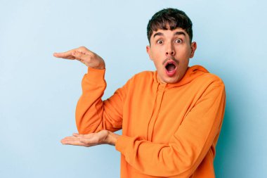 Genç melez adam mavi arka planda izole edilmiş şok olmuş ve hayrete düşmüş bir şekilde elleri arasında bir kopya alanı tutuyor..