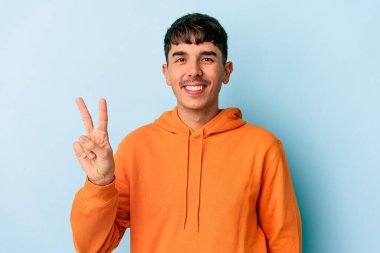 Genç melez adam mavi arka planda izole edilmiş zafer işareti gösteriyor ve gülümsüyor..