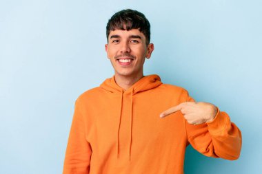 Genç melez adam mavi arka planda izole edildi. Elle bir tişört fotokopi alanını işaret ediyor. Gururlu ve kendinden emin.