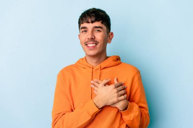 Mavi arka planda izole edilmiş genç melez adam avuçlarını göğsüne bastıran dostane bir ifadeye sahiptir. Aşk konsepti.