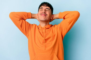 Genç melez adam mavi arka planda izole edilmiş. Kendine güveniyor, elleri başının arkasında..