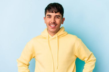 Genç melez adam mavi arka planda izole edilmiş. Kendine güvenen, ellerini kalçasında tutan..