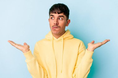 Genç melez adam mavi arka planda izole edilmiş. Kafası karışmış ve omuz silkiyor. Kopya alanını tutmak için..