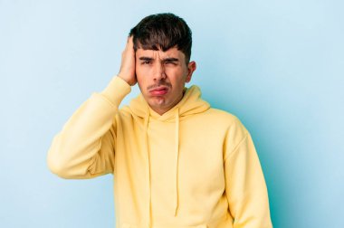 Genç melez adam mavi arka planda izole edilmiş. Çok yorgun ve uykulu. Elini başının üstünde tutuyor..