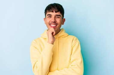 Genç melez adam mavi arka planda izole edilmiş mutlu ve kendinden emin gülümsüyor, elleriyle çenesine dokunuyor..