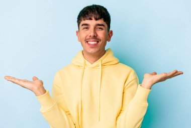 Mavi arka planda soyutlanmış genç melez adam kollarıyla ölçeği yapıyor, mutlu ve kendinden emin hissediyor..