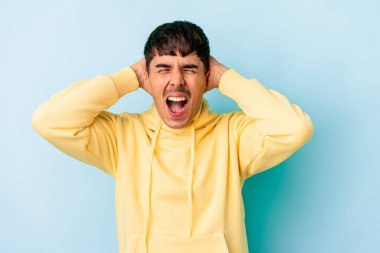 Genç melez adam mavi arka planda izole edilmiş kulakları kulaklarıyla kapatıyor çok fazla ses duymamaya çalışıyor..
