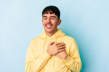 Genç melez adam mavi arka planda soyutlanmış gülüyor, ellerini kalbe koyuyor, mutluluk kavramını koruyor..