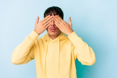 Genç melez adam mavi arka planda izole edilmiş elleri olan gözleri kapatmaktan korkuyor..