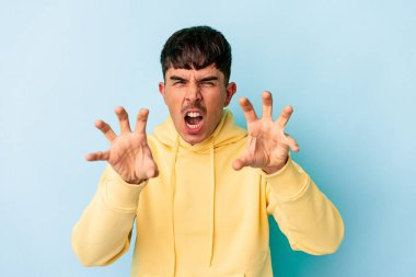 Mavi arka planda izole edilmiş genç melez bir adam pençelerini kedi taklidi yaparken gösteriyor, agresif bir hareket..