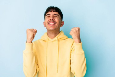 Genç melez adam mavi arka planda izole edilmiş zaferi, tutkuyu ve coşkuyu kutluyor, mutlu bir ifade..