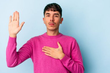 Genç melez adam mavi arka planda izole edilmiş yemin ediyor, elini göğsüne koyuyor..