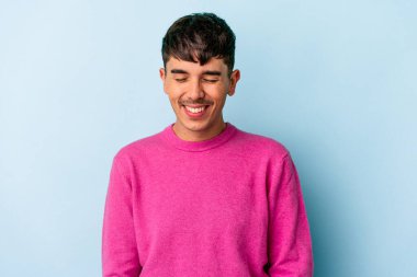 Genç melez adam mavi arka planda izole edilmiş gülüyor ve gözlerini kapıyor, rahatlamış ve mutlu hissediyor..