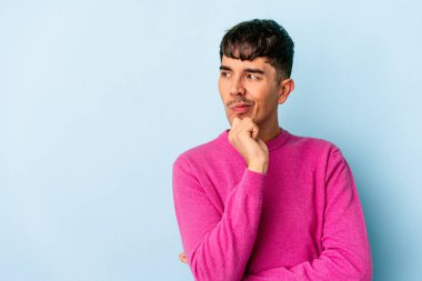 Genç melez adam mavi arka planda izole edilmiş şüpheli ve şüpheci bir ifadeyle yana bakıyor..