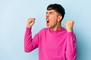 Genç melez adam mavi arka planda izole edilmiş zafer sonrası yumruğunu kaldırıyor, kazanan konsept..