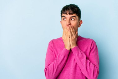 Genç melez adam mavi arka planda izole edilmiş. Düşünceli bir şekilde bir fotokopi odasına bakıyor. Ağzını eliyle kapatıyor..