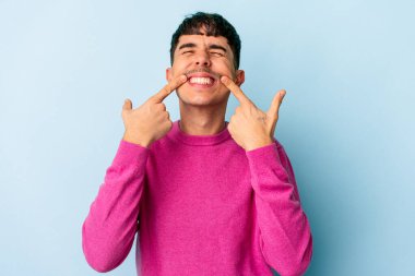 Genç melez adam mavi arka planda izole edilmiş gülümsüyor, parmaklarını ağıza doğrultuyor..