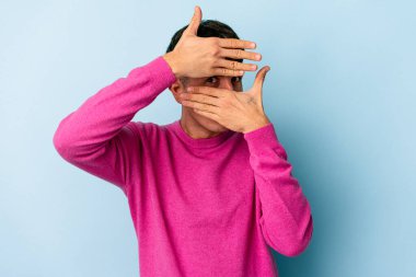 Genç melez adam mavi arka planda izole edilmiş parmaklarıyla kameraya göz kırpıyor, yüzünü kapatmaktan utanıyor..