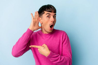 Genç melez adam mavi arka planda izole edilmiş bir şekilde bir dedikoduyu dinlemeye çalışıyor..