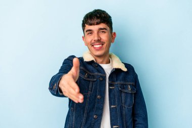 Genç melez adam mavi arka planda izole edilmiş kameraya elini uzatarak selam veriyor..