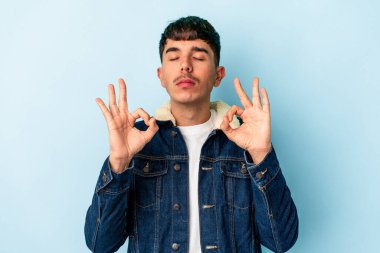 Genç melez adam mavi arka planda izole edilmiş yoğun bir günün ardından rahatlıyor, yoga yapıyor..