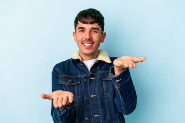 Mavi arka planda soyutlanmış genç melez adam kollarıyla ölçeği yapıyor, mutlu ve kendinden emin hissediyor..