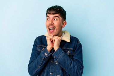 Mavi arka planda izole genç melez adam ellerini çenesinin altında tutuyor, mutlu bir şekilde yana bakıyor..