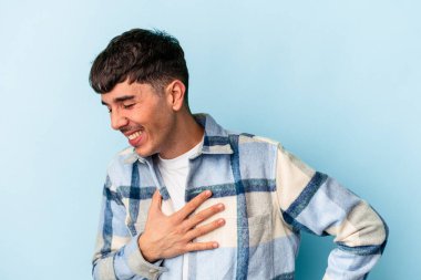 Genç melez adam mavi arka planda soyutlanmış gülüyor, ellerini kalbe koyuyor, mutluluk kavramını koruyor..