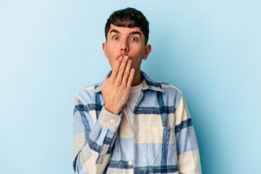 Genç melez adam mavi arka planda izole edilmiş şok olmuş, ağzı ellerle kaplanmış, yeni bir şey keşfetmeye hevesli..