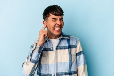 Genç melez adam mavi arka planda izole edilmiş kulakları parmaklarıyla kapatıyor, stresli ve çaresiz bir ortam tarafından..