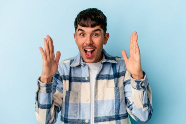 Genç melez adam mavi arka planda izole edilmiş. Hoş bir sürpriz bekliyor, heyecanlı ve el kaldırıyor..