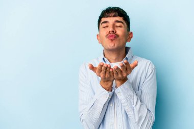 Genç melez adam mavi arka planda izole edilmiş dudaklar kıvrılıyor ve hava öpücüğü göndermek için avuç içi tutuyor..