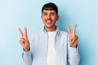 Genç melez adam mavi arka planda izole edilmiş zafer işareti gösteriyor ve gülümsüyor..