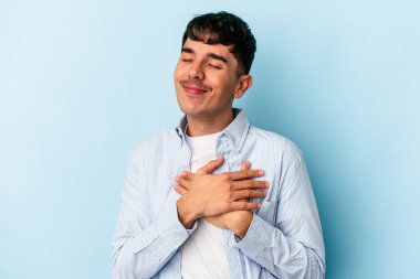 Mavi arka planda izole edilmiş genç melez adam avuçlarını göğsüne bastıran dostane bir ifadeye sahiptir. Aşk konsepti.