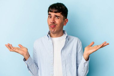 Genç melez adam, mavi arka planda izole edilmiş sorgulama jestinde şüphe ediyor ve omuz silkiyor..