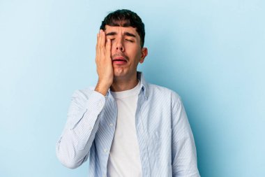 Genç melez adam mavi arka planda izole edilmiş. Çok yorgun ve uykulu. Elini başının üstünde tutuyor..