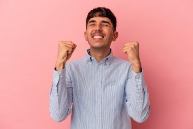 Genç melez adam pembe arka planda izole edilmiş zaferi, tutkuyu ve coşkuyu kutluyor, mutlu bir ifade..