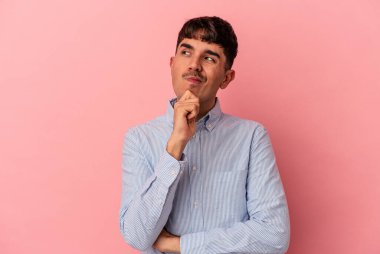 Genç melez adam pembe arka planda izole edilmiş düşünüyor, bakıyor, yansıtıyor, düşünüyor, hayal kuruyor..