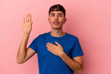 Genç melez adam pembe arka planda izole edilmiş yemin ediyor, elini göğsüne koyuyor..