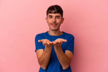 Genç melez adam pembe arka planda izole edilmiş. Elinde avuç içi olan bir şeyle kameraya sunuyor..