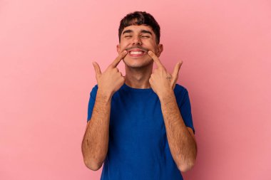 Genç melez adam pembe arka planda izole edilmiş gülümsüyor, parmaklarını ağıza doğrultuyor..